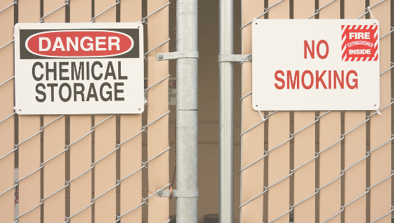 Decorative image of gate with two safety signs. One reads "Caution Chemical Storage" and the other reads "No Smoking"